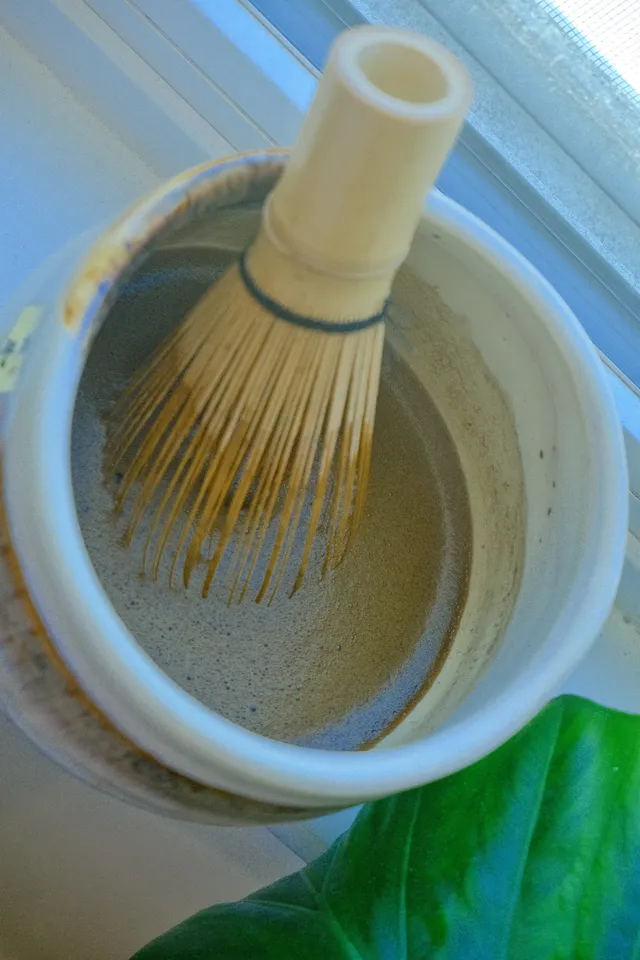 Bamboo whisk dipped into a bowl of freshly whisked hojicha, foam settling near a window